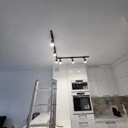 Mateusz Myśków Usługi Elektryczne - Montaż oświetlenia szynowego w nowoczesnej kuchni z białymi meblami. Widoczna drabina i lampy sufitowe.