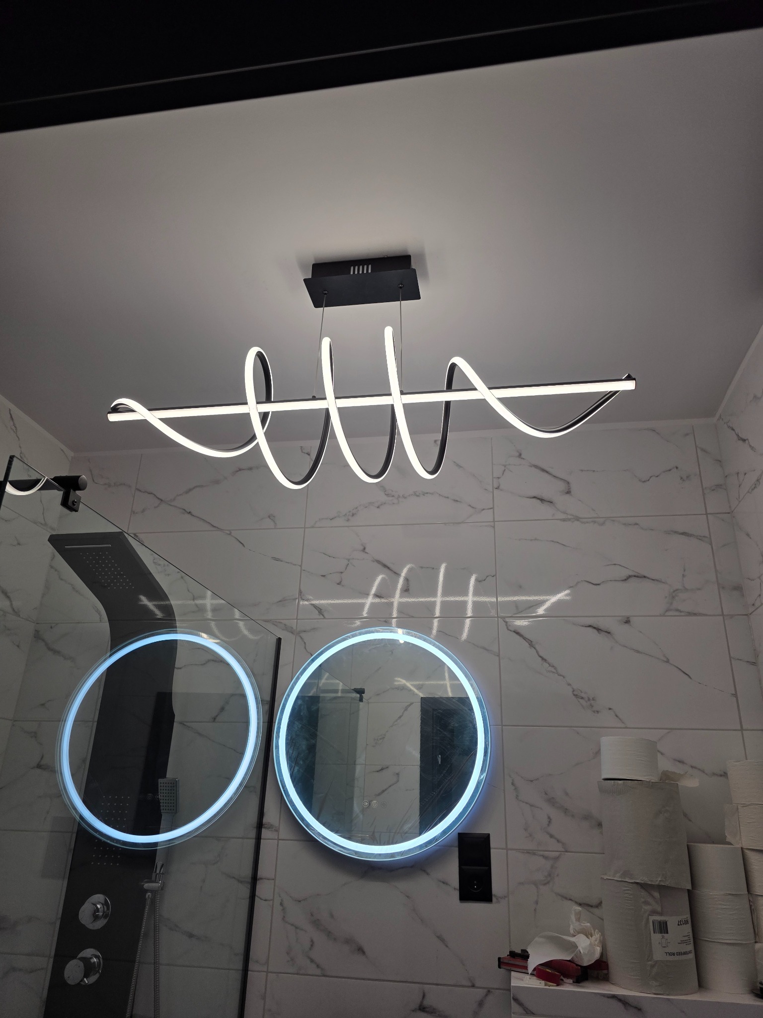 Nowoczesna lampa LED w kształcie spirali, zamontowana w łazience z marmurowymi płytkami i okrągłymi lustrami z podświetleniem. Widoczna kabina prysznicowa.