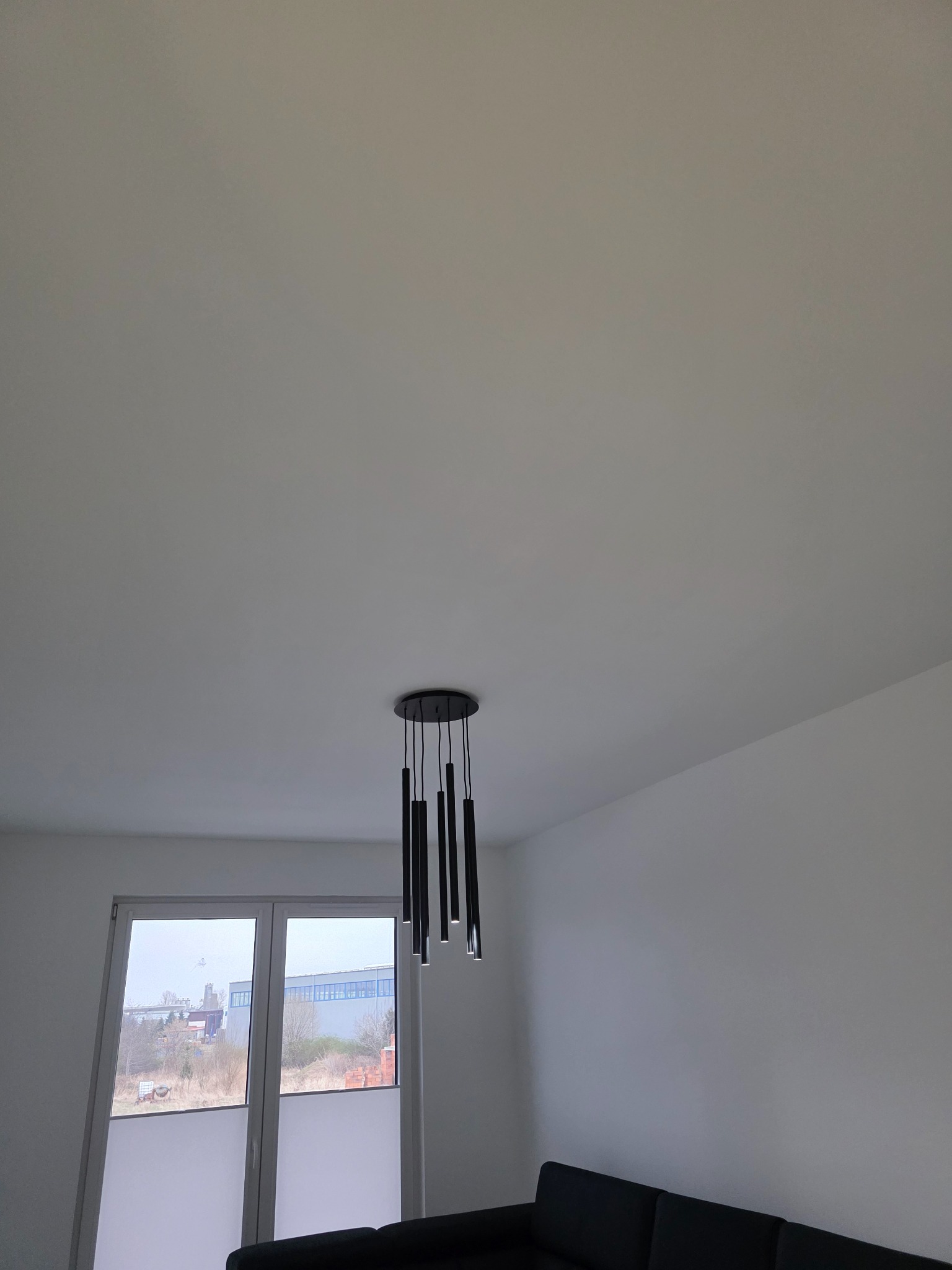 Czarny, nowoczesny żyrandol z podłużnymi lampami w Wałbrzychu, zamontowany na białym suficie. W tle okno z widokiem na zewnątrz i fragment czarnej sofy.