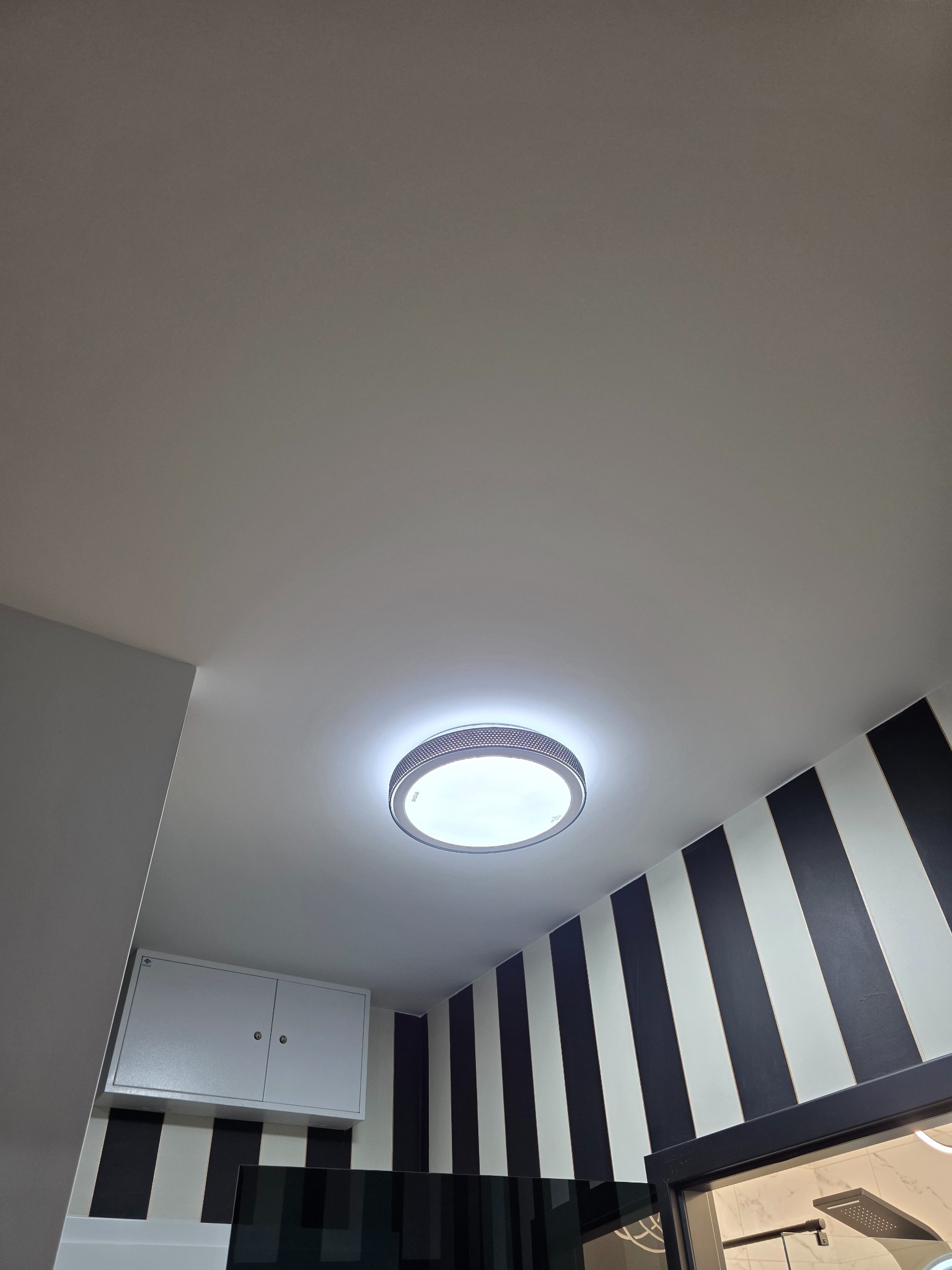 Zamontowana okrągła lampa sufitowa LED w łazience z czarno-białymi pasami na ścianie. Widok z dołu, oświetlenie jasne, nowoczesny design.