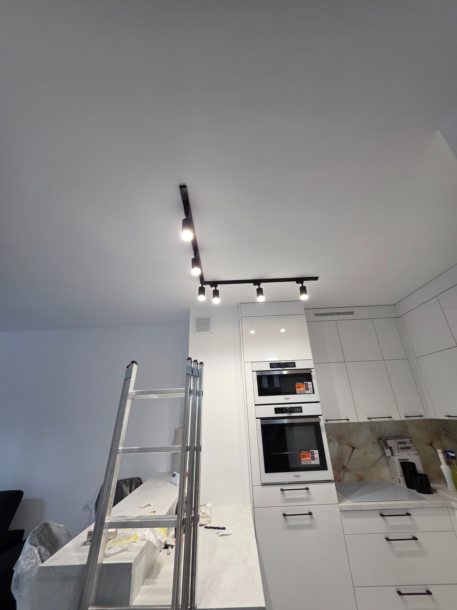 Montaż oświetlenia szynowego w nowoczesnej kuchni z białymi meblami. Widoczna drabina i lampy sufitowe.
