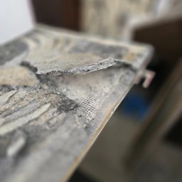 Agnieszka Czajkowska - Odkruszony fragment ściany z siatką wzmacniającą, ukazujący strukturę i warstwy materiału. Nierówna powierzchnia, bliskie ujęcie z rozmytym tłem.