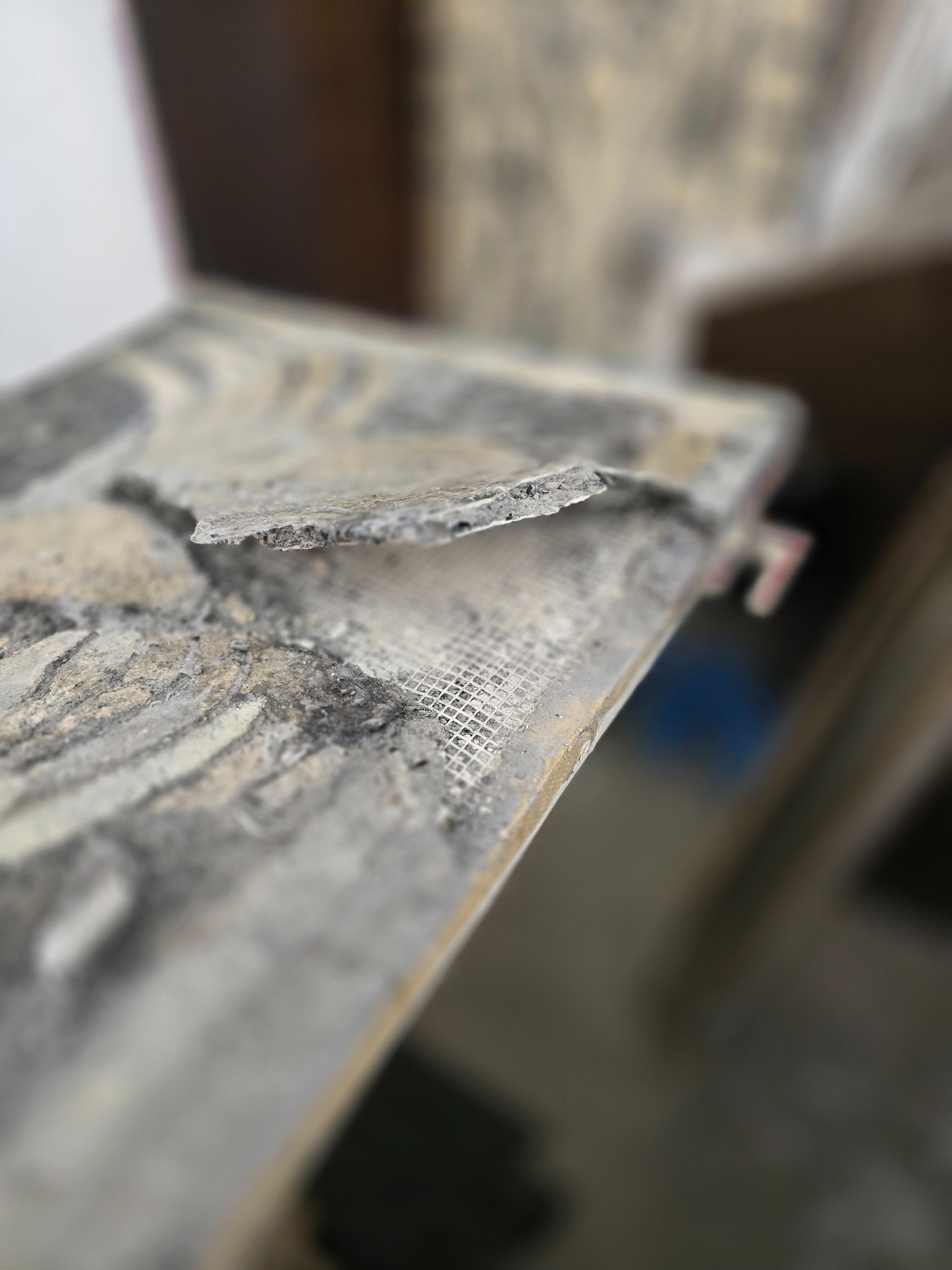 Odkruszony fragment ściany z siatką wzmacniającą, ukazujący strukturę i warstwy materiału. Nierówna powierzchnia, bliskie ujęcie z rozmytym tłem.