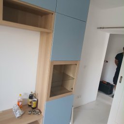 Piotr Jockel - Narożna zabudowa meblowa w odcieniach błękitu i drewna, z przeszkloną witryną, na tle białej ściany i marmurowej podłogi. Widoczne akcesoria i narzędzia.