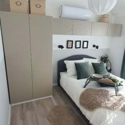 Piotr Jockel - Komfortowa sypialnia z beżową szafą, łóżkiem z tacą śniadaniową i dywanikiem. Na ścianie dekoracyjne ramki i klimatyzator. Jasne panele podłogowe.