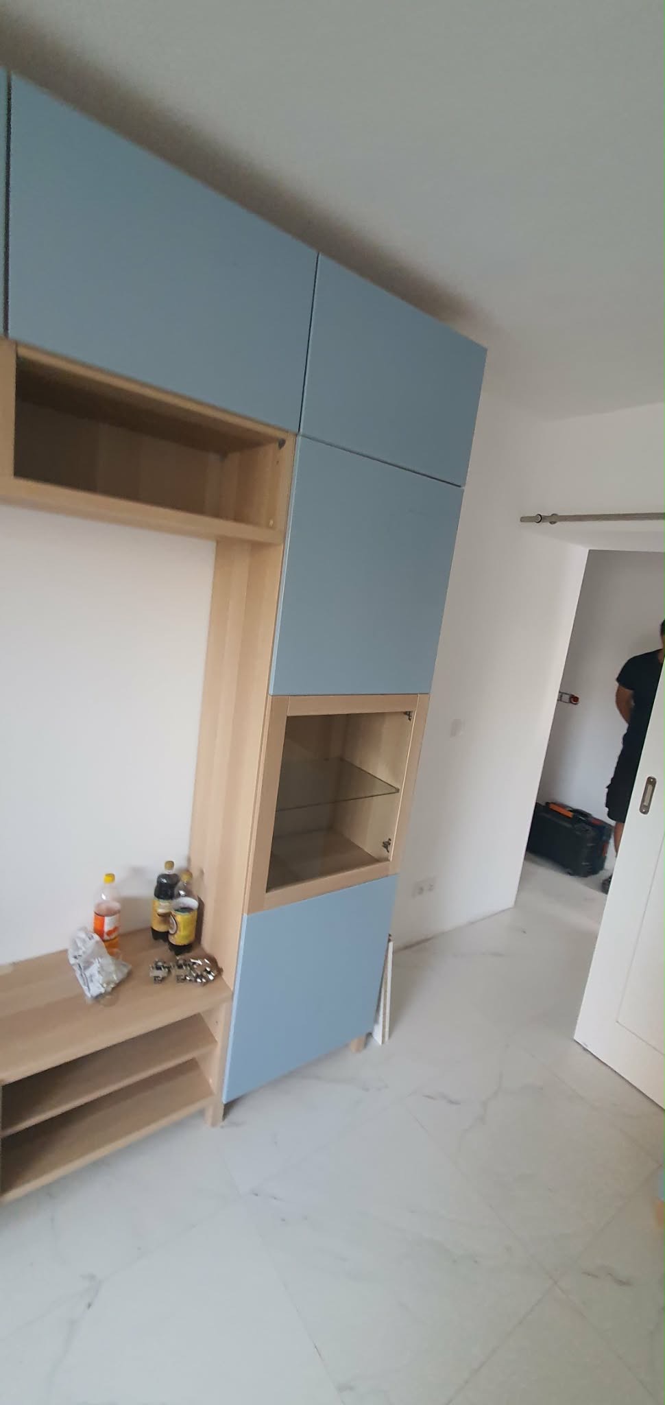 Narożna zabudowa meblowa w odcieniach błękitu i drewna, z przeszkloną witryną, na tle białej ściany i marmurowej podłogi. Widoczne akcesoria i narzędzia.