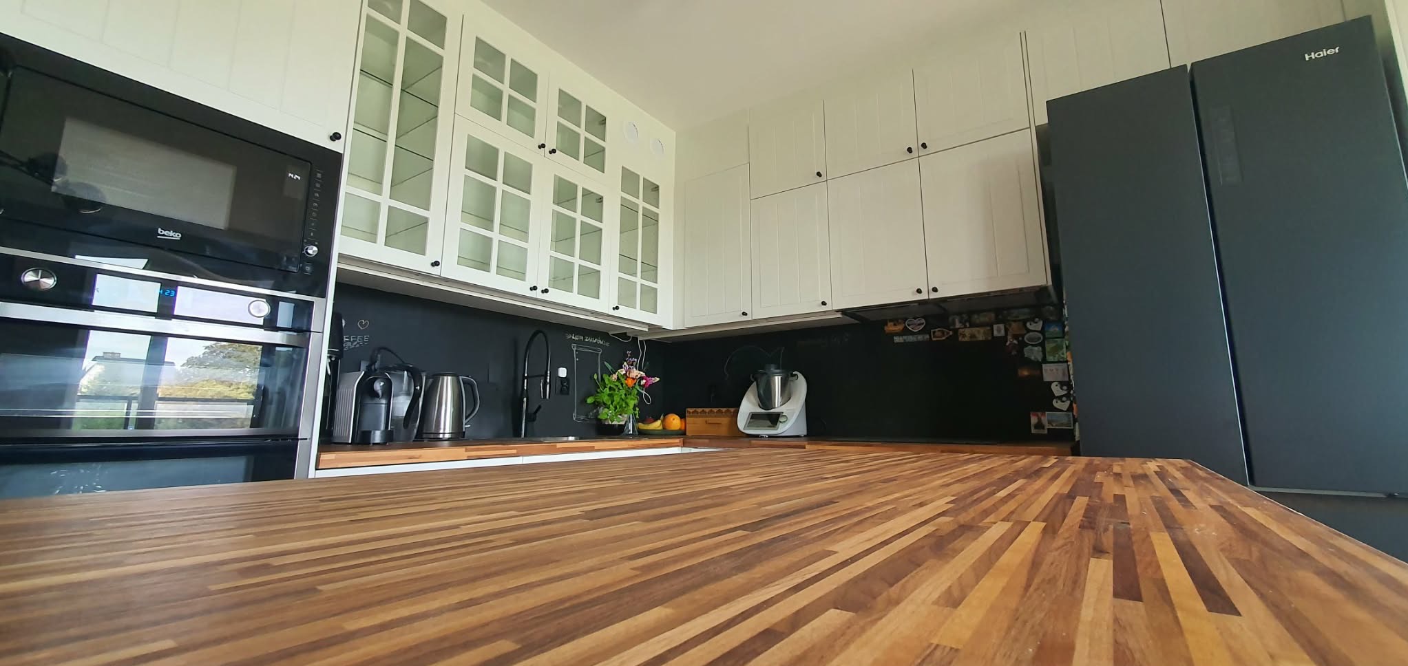 Nowoczesna kuchnia z białymi szafkami, czarnym blatem i sprzętem AGD. Uwagę przykuwa drewniany blat wyspy kuchennej z wyraźnym usłojeniem.