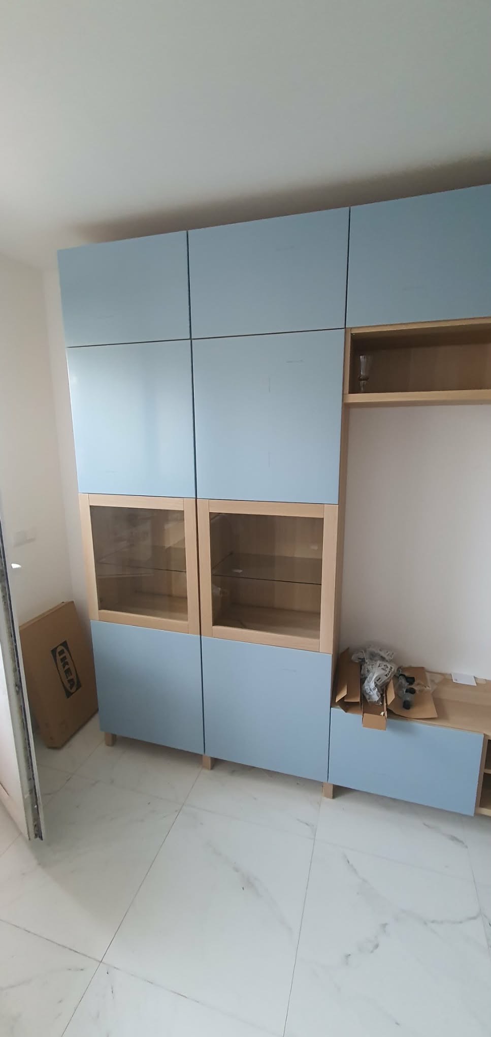 Niebiesko-drewniany regał z przeszklonymi witrynami, częściowo zmontowany, z kartonem IKEA obok. Widok na marmurowe płytki podłogowe.