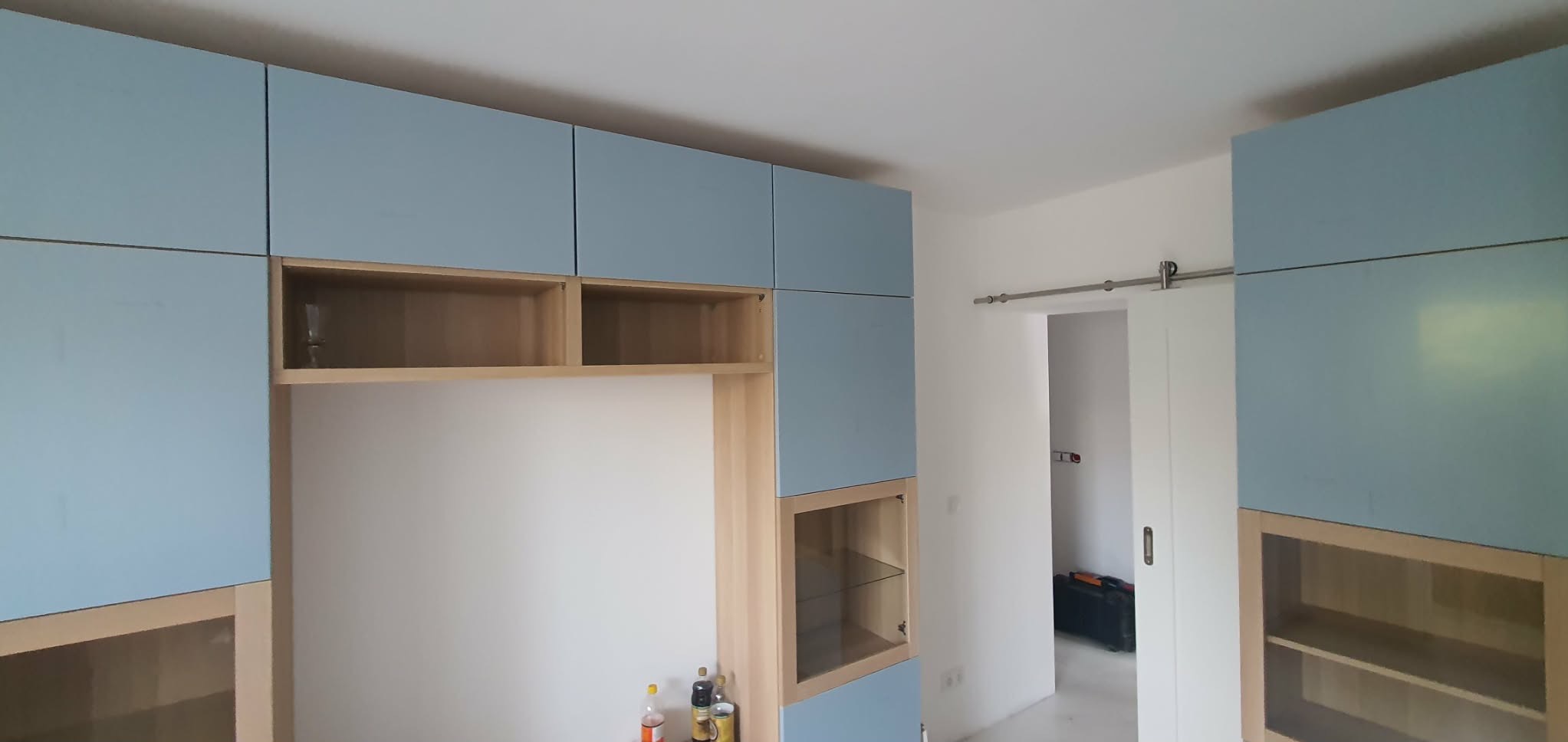 Niebiesko-drewniana zabudowa meblowa w salonie, widok z dołu. Częściowo zamontowane szafki wiszące, przesuwne drzwi do innego pomieszczenia w tle.