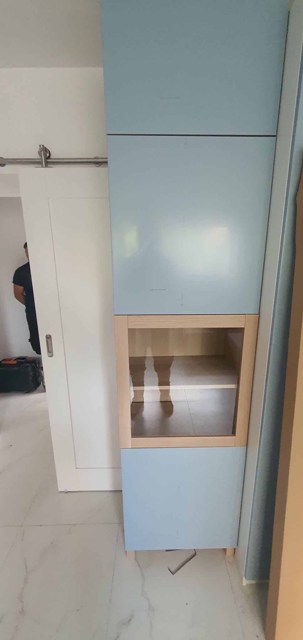 Niebieski regał z drewnianą wnęką, widoczny na tle białych drzwi przesuwnych i marmurowej podłogi. W wnęce odbija się sylwetka osoby.