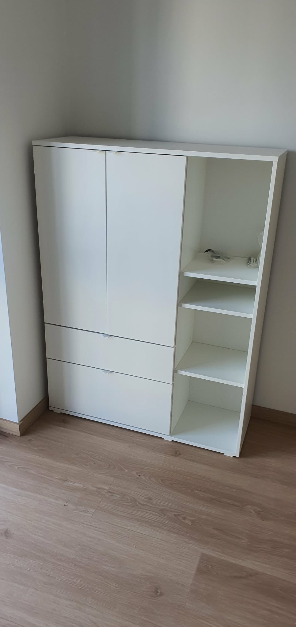 Biała komoda z dwiema szafkami i trzema szufladami, z otwartą półką po prawej stronie, stoi na panelach podłogowych w jasnym odcieniu drewna.