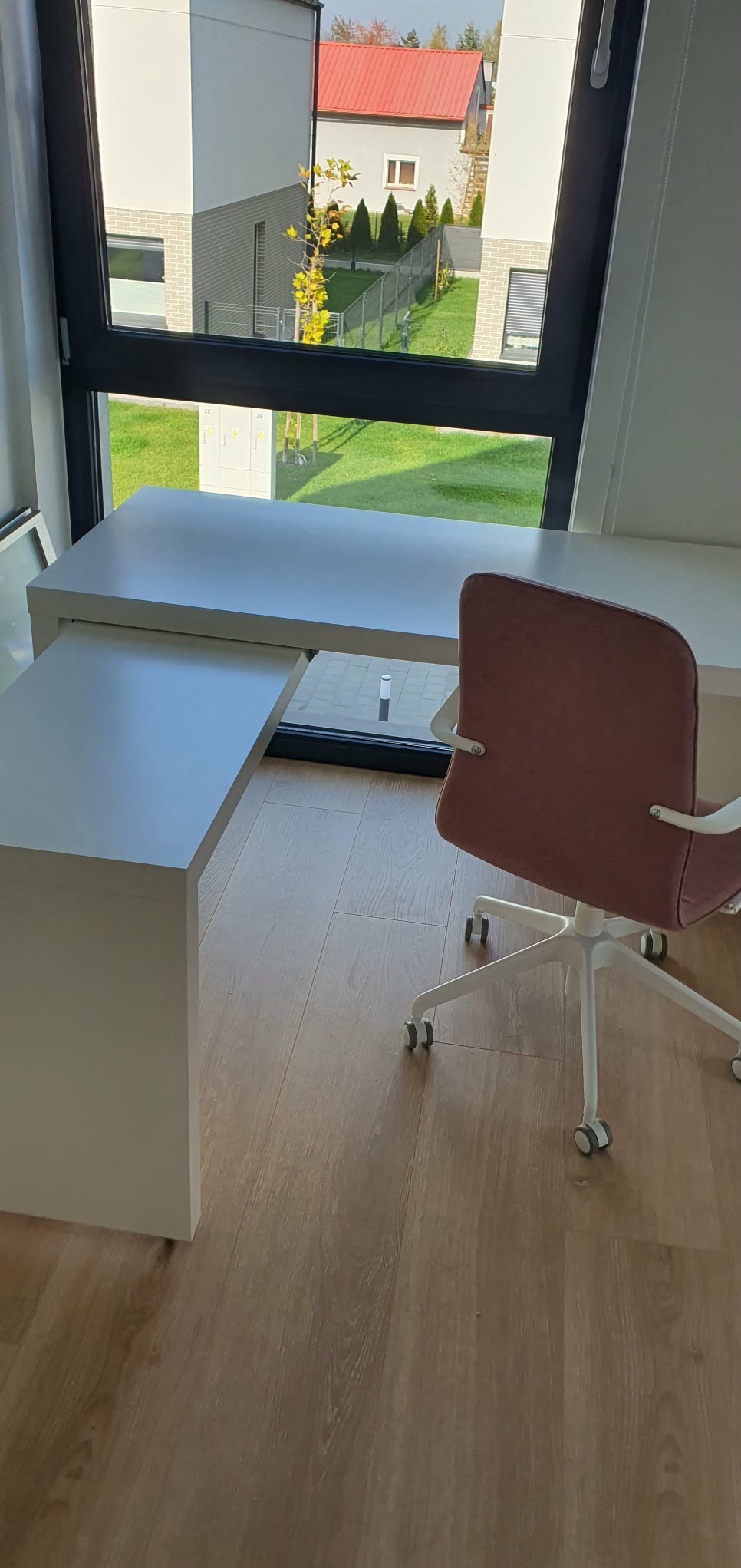 Białe biurko z wysuwanym blatem i różowe krzesło biurowe na drewnianej podłodze, widok z okna na budynki i zieleń.
