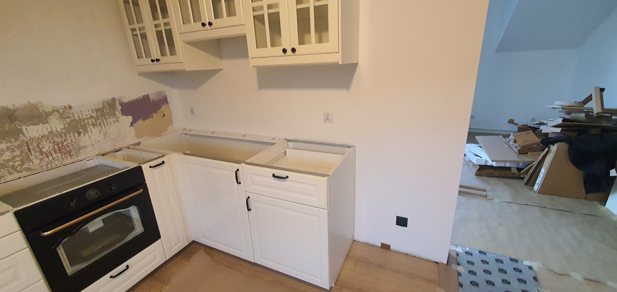 Wnętrze kuchni w trakcie prac: białe szafki dolne i wiszące, ściany częściowo przygotowane do malowania, widoczne kartony i materiały budowlane w tle.