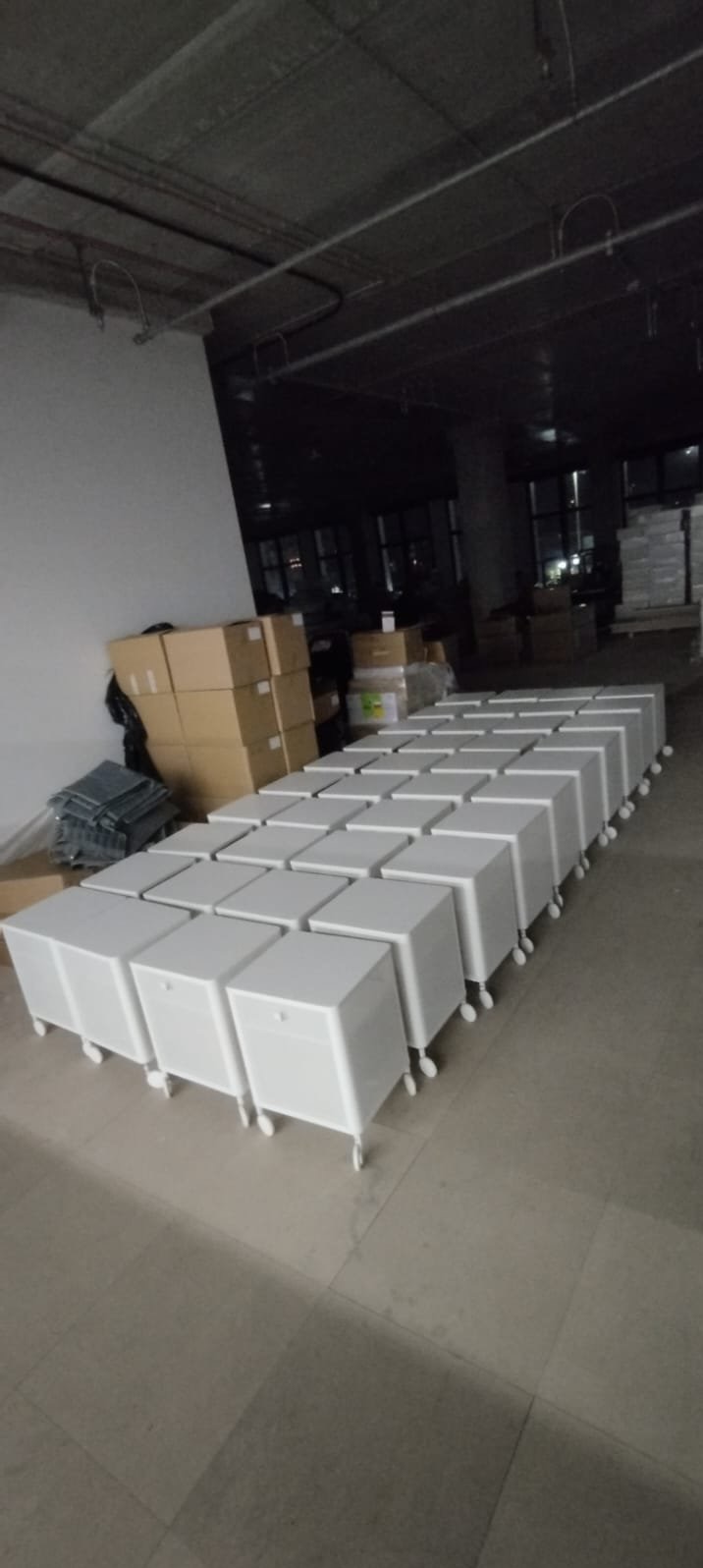 Rząd białych szafek na kółkach, z szufladami i uchwytami, ustawionych w rzędach w surowym wnętrzu. W tle kartony i elementy montażowe.