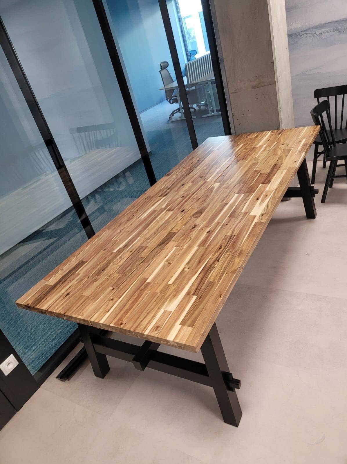 Długi, drewniany stół z blatem z klejonego drewna i czarnymi nogami, ustawiony w nowoczesnym biurze z przeszkloną ścianą. Widoczne krzesła w tle.