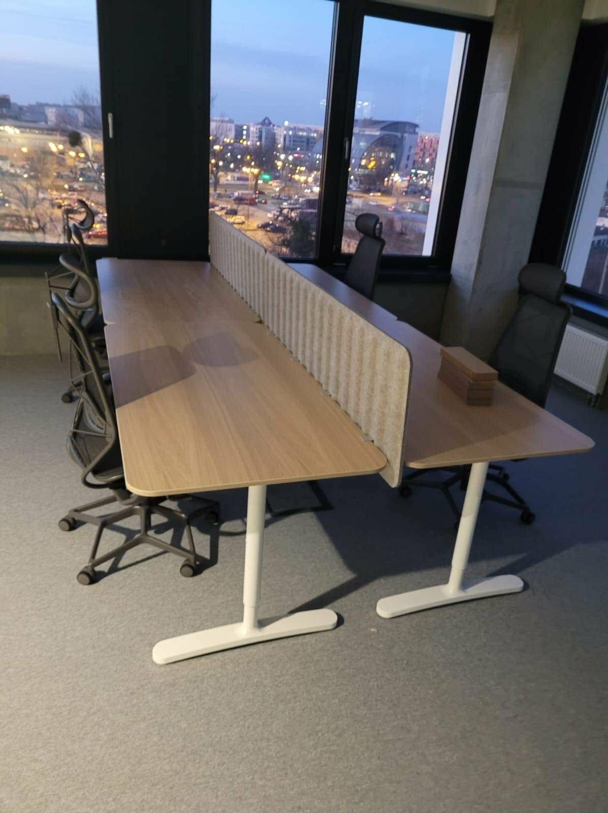 Długi, jasny biurkowy stół z przegrodami w nowoczesnym biurze. Widok na miasto przez okno. Krzesła biurowe w tle. Minimalistyczny design.