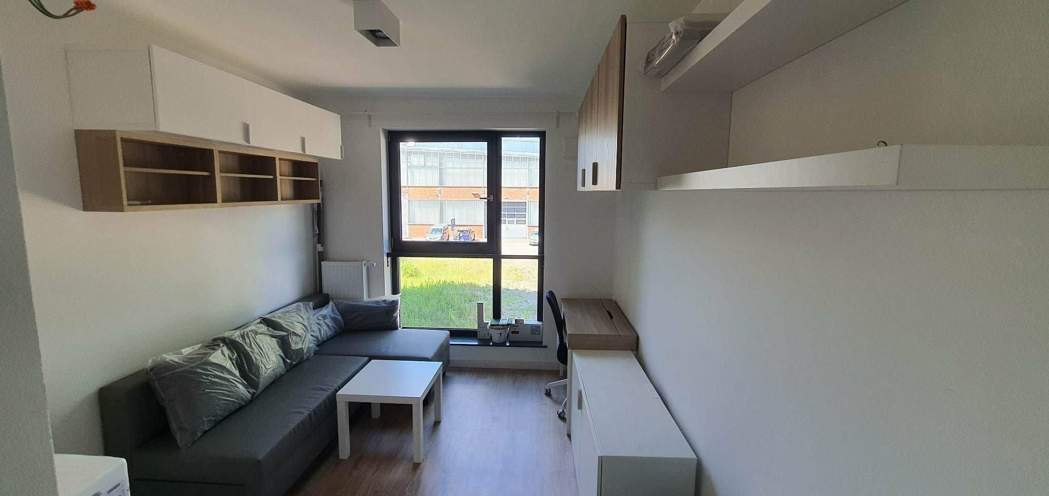 Nowoczesny pokój z szarą sofą, białym stolikiem i biurkiem przy oknie. Na ścianach wiszące szafki i półki. Widok na zewnątrz przez duże okno.