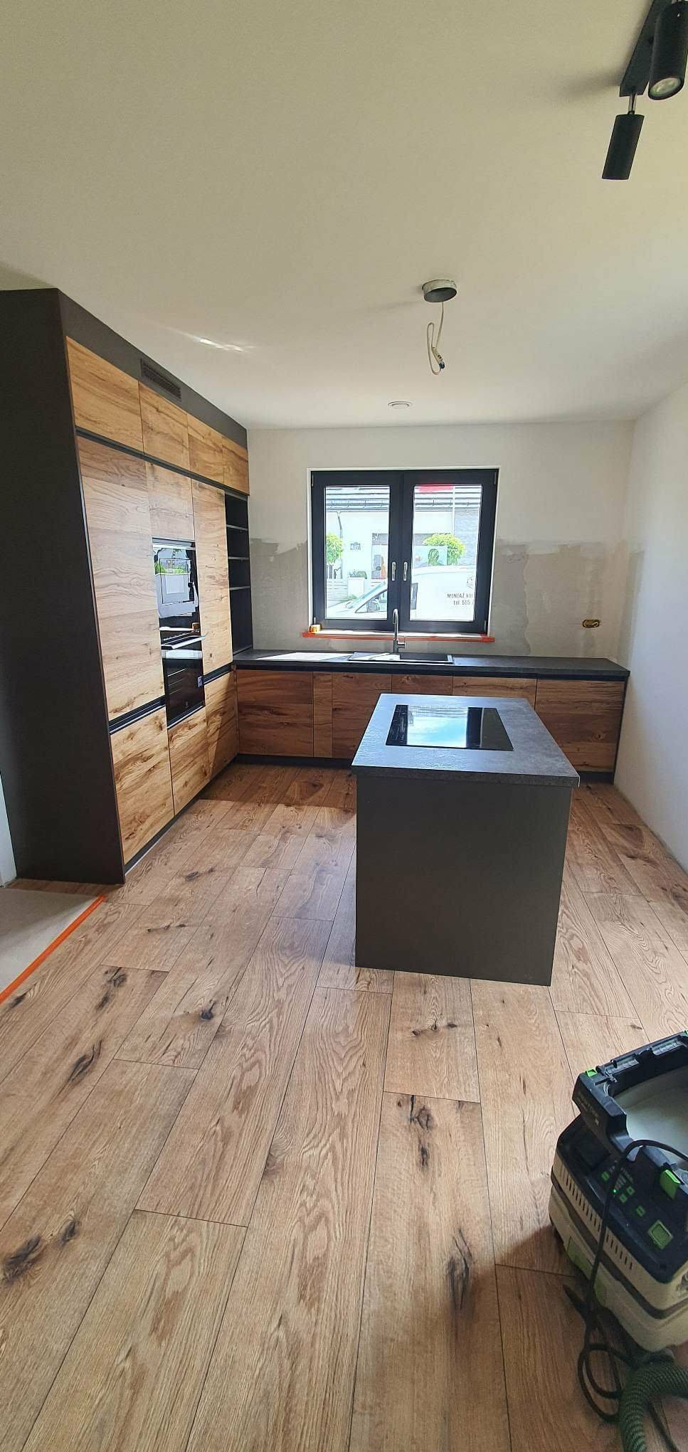 Nowoczesna kuchnia z drewnianymi frontami i wyspą, widok na okno i parkiet. Częściowo wykończone ściany, narzędzie na podłodze sugeruje trwające prace.