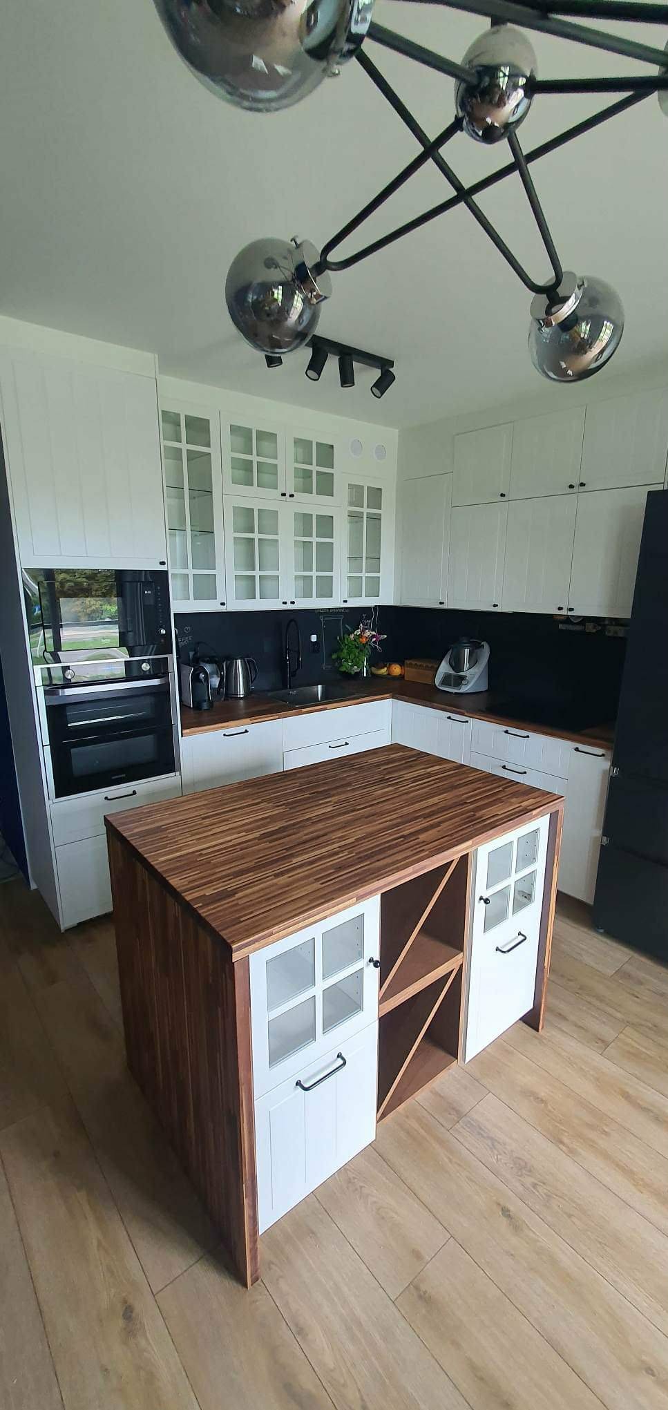 Nowoczesna kuchnia z wyspą o drewnianym blacie i białych frontach. Widoczne białe szafki, czarny panel nad blatem i designerska lampa sufitowa.