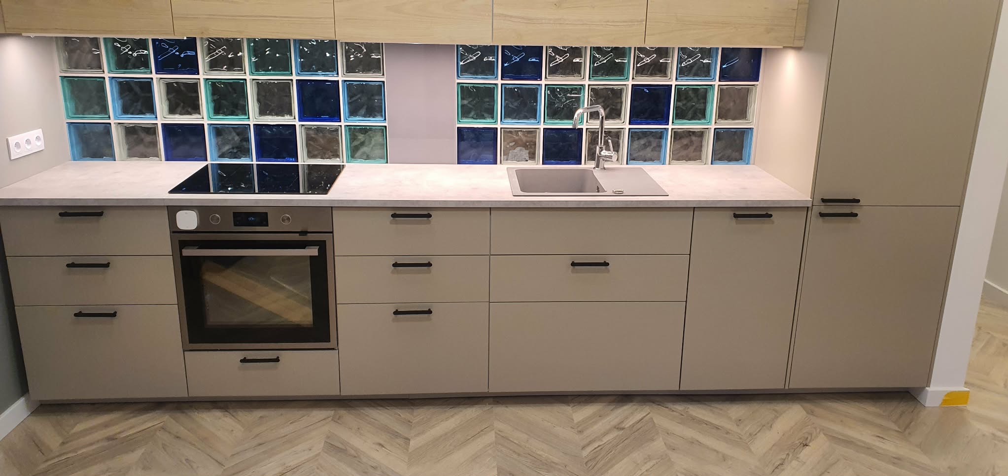 Beżowa zabudowa kuchenna z czarnymi uchwytami, szklane kafle w odcieniach niebieskiego i zielonego nad blatem, płyta indukcyjna i piekarnik wkomponowane w meble.