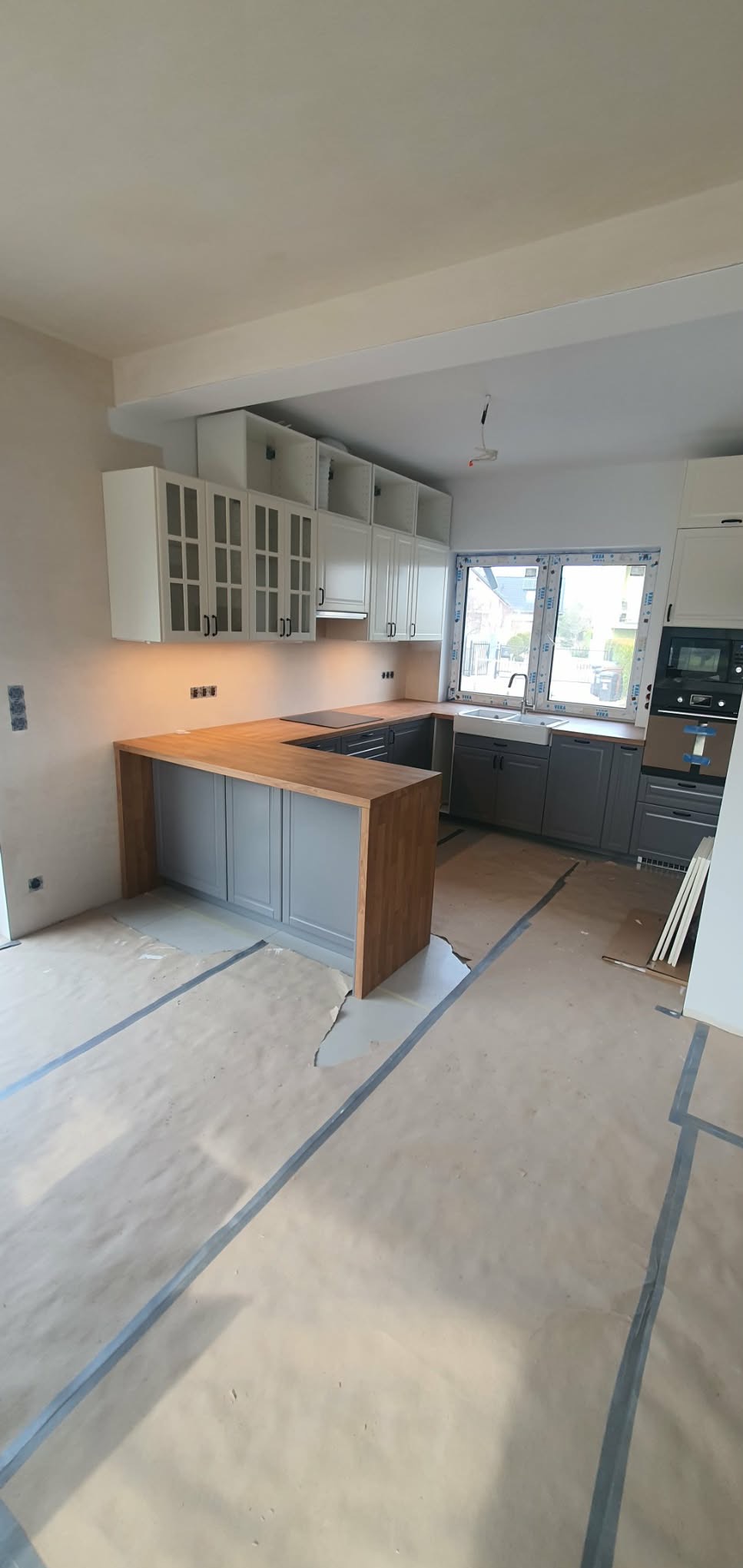 Jasna kuchnia z drewnianym blatem i szarymi szafkami, widok na okno i częściowo zabezpieczoną podłogę. Górne szafki białe, niektóre przeszklone.