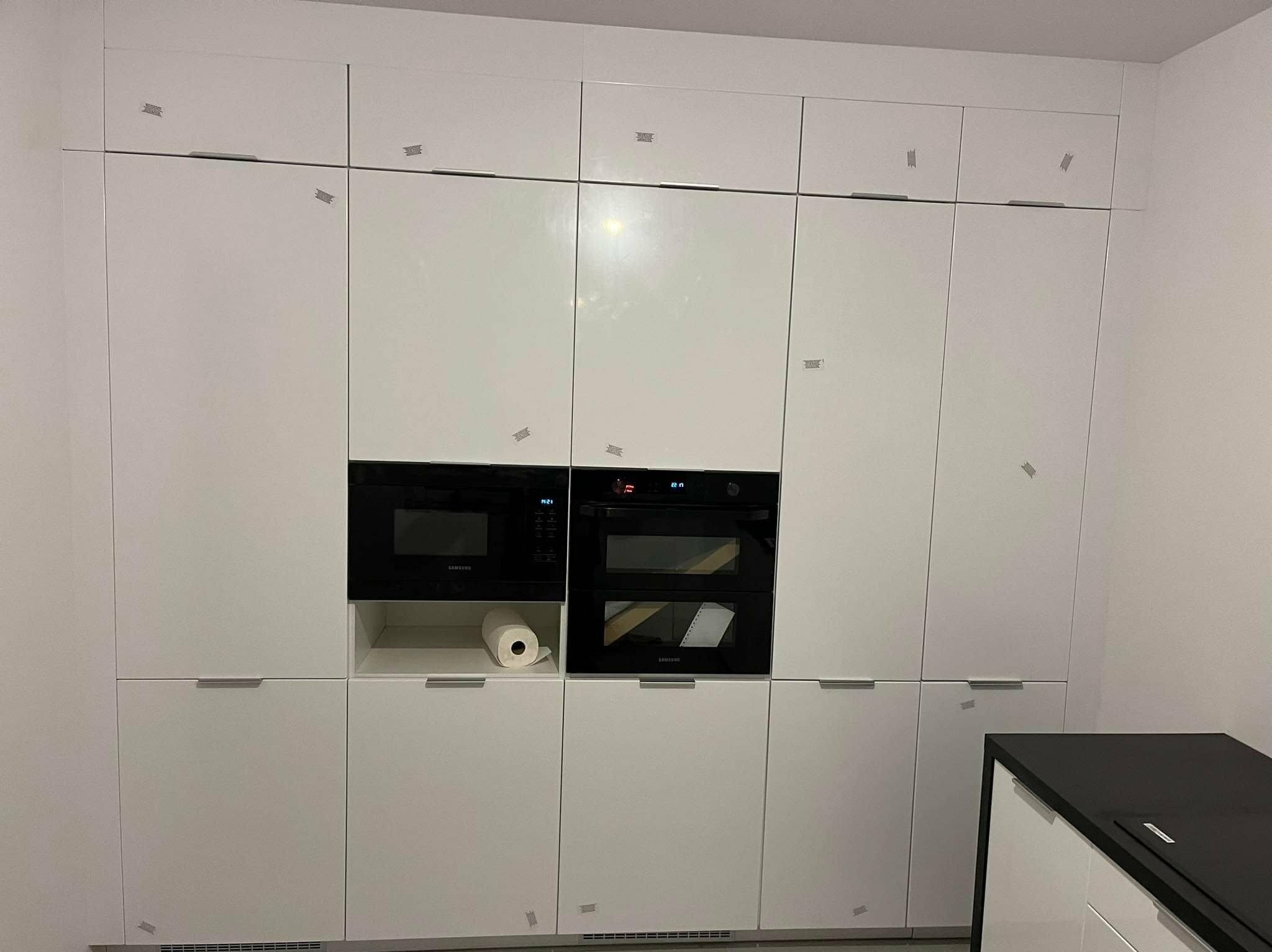 Biała zabudowa kuchenna z piekarnikiem i mikrofalówką Samsung, zabezpieczona taśmą malarską. Minimalistyczny design, nowoczesne wykończenie.
