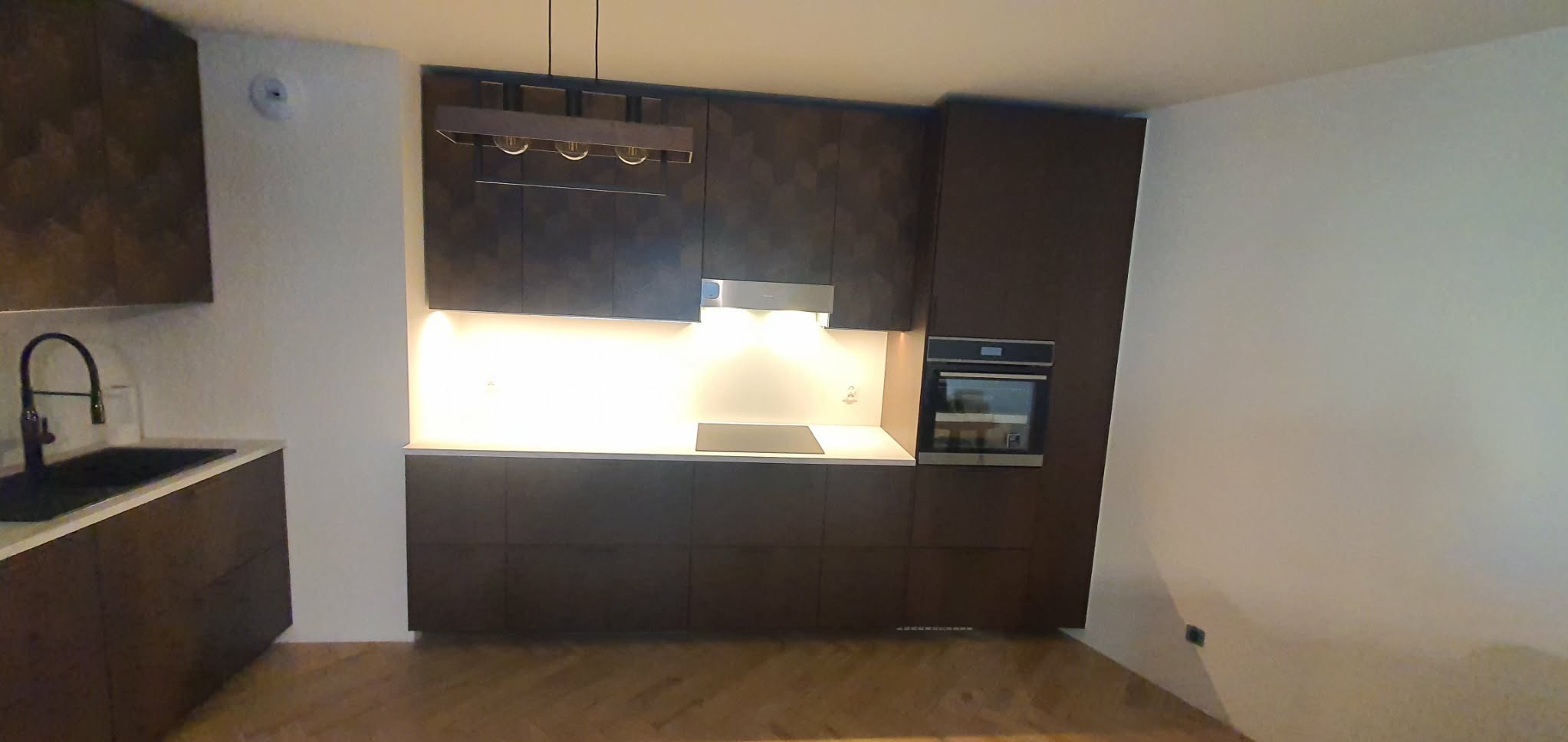 Nowoczesna kuchnia z ciemnymi, geometrycznymi frontami szafek, białym blatem i oświetleniem LED pod szafkami. Widoczny piekarnik i płyta indukcyjna. Parkiet na podłodze.