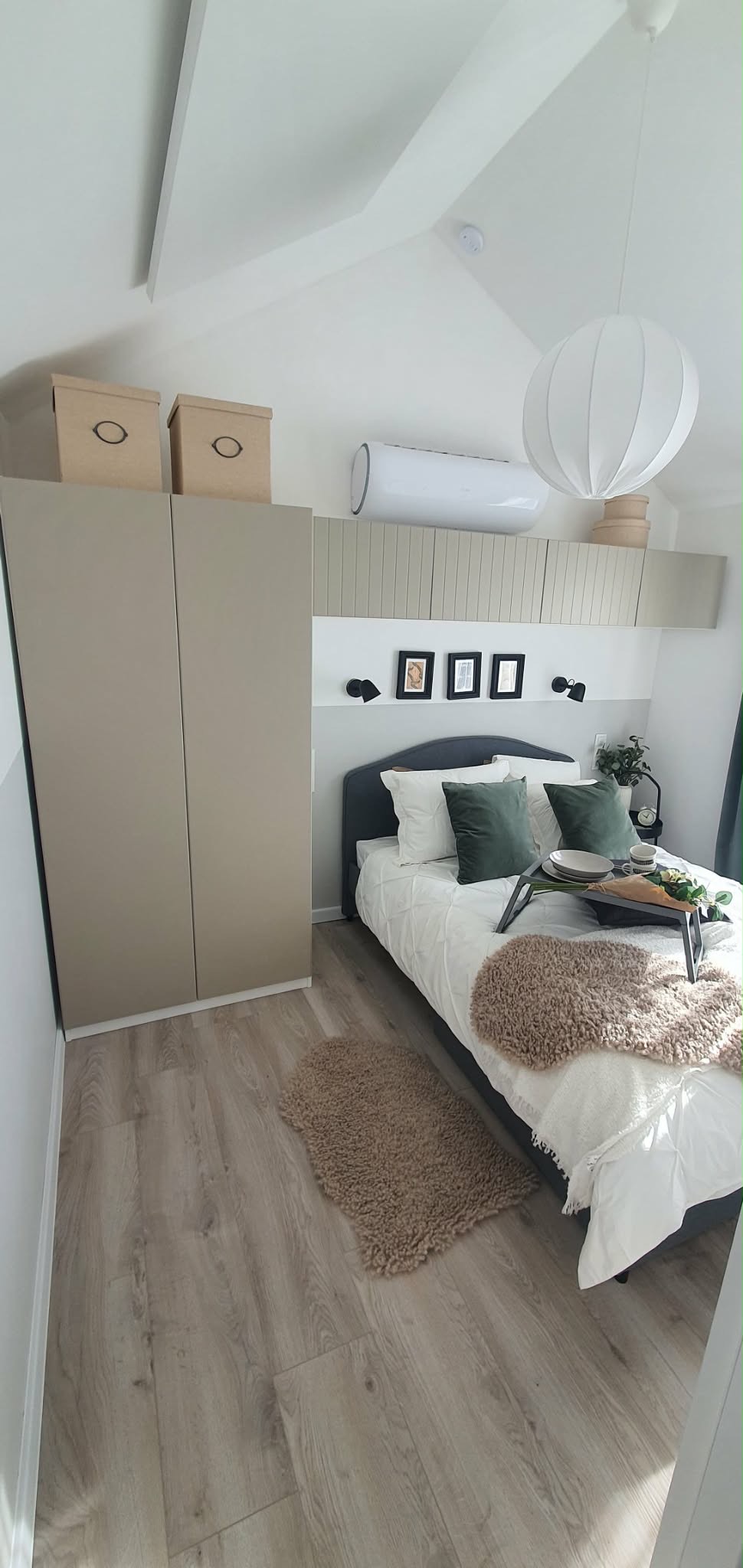 Komfortowa sypialnia z beżową szafą, łóżkiem z tacą śniadaniową i dywanikiem. Na ścianie dekoracyjne ramki i klimatyzator. Jasne panele podłogowe.