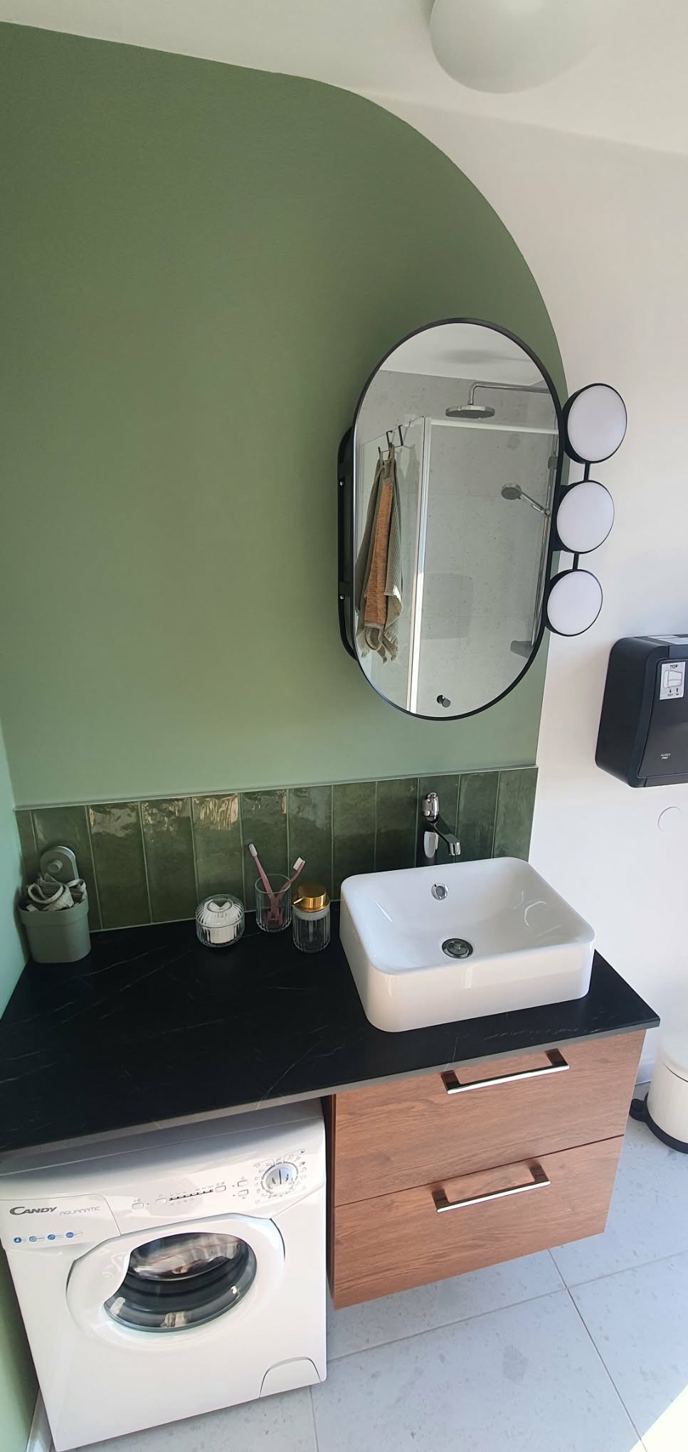 Nowoczesna łazienka z umywalką nablatową, lustrem z oświetleniem i pralką Candy. Ściana w kolorze oliwkowym, płytki w odcieniach zieleni. Minimalistyczny design.