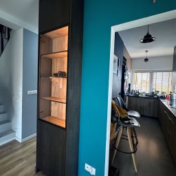 Partner Producent Mebli Na Wymiar - Ciemna, wysoka witryna z podświetlanymi półkami, eksponująca kolekcję modeli aut, w nowoczesnym wnętrzu z turkusową ścianą i widokiem na kuchnię.