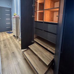 Partner Producent Mebli Na Wymiar - Otwarta szafa wnękowa z oświetleniem LED, drewnianymi półkami i szufladami w odcieniach szarości. Widoczne wysuwane półki i fragment przedpokoju z komodą.