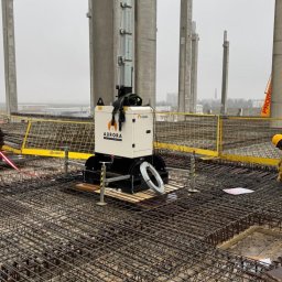 Elchin Japarov - Zbrojenie płyty fundamentowej z widoczną siatką zbrojeniową, słupami konstrukcyjnymi i przenośną wieżą oświetleniową Aurora na budowie w pochmurny dzień.