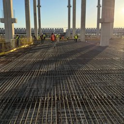 Elchin Japarov - Siatka zbrojeniowa na budowie, widok z góry, betonowe kolumny w tle, pracownicy w oddali, wczesne popołudnie, delikatne światło słoneczne.