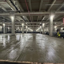Elchin Japarov - Świeżo wylana, mokra posadzka w hali z betonowymi kolumnami i rurami pod sufitem. W tle widoczni pracownicy w odblaskowych ubraniach i maszyna do wylewania betonu.