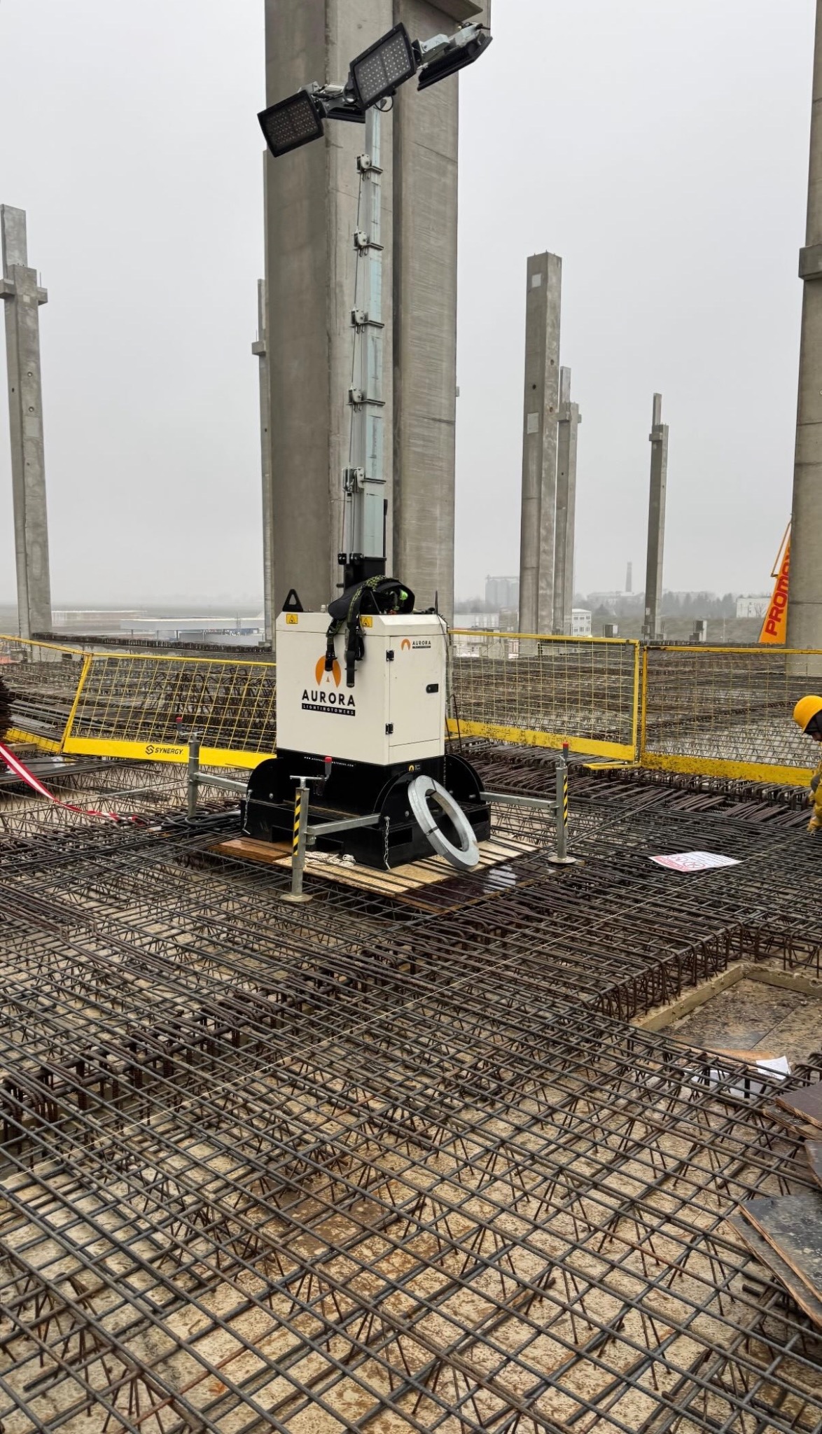 Zbrojenie płyty fundamentowej z widoczną siatką zbrojeniową, słupami konstrukcyjnymi i przenośną wieżą oświetleniową Aurora na budowie w pochmurny dzień.
