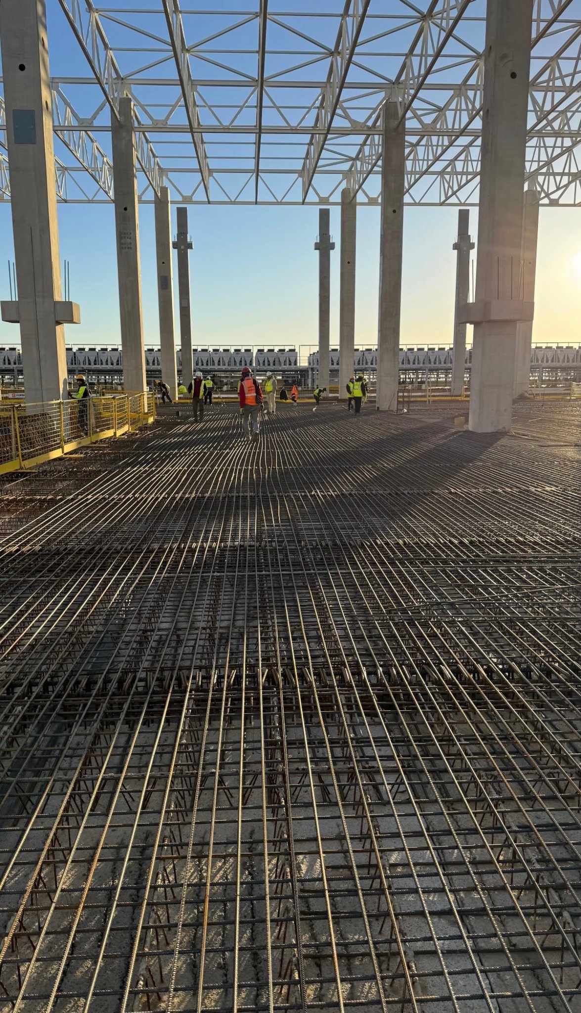 Siatka zbrojeniowa na budowie, widok z góry, betonowe kolumny w tle, pracownicy w oddali, wczesne popołudnie, delikatne światło słoneczne.