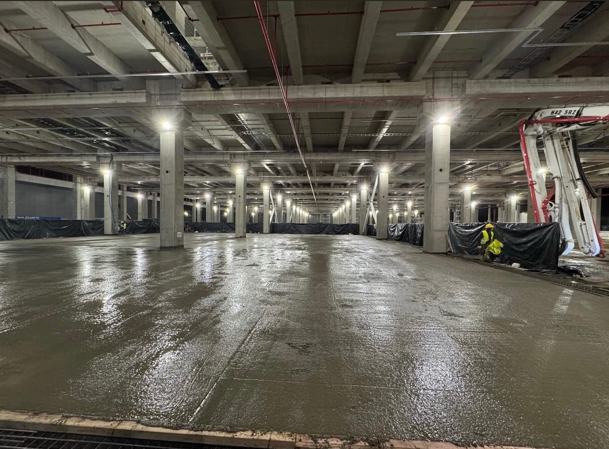 Świeżo wylana, mokra posadzka w hali z betonowymi kolumnami i rurami pod sufitem. W tle widoczni pracownicy w odblaskowych ubraniach i maszyna do wylewania betonu.