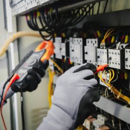 J&M Professional - Elektryk w szarych rękawicach z czarnymi dłońmi diagnozuje szafę elektryczną, mierząc prąd cęgowym miernikiem i napięcie sondą na elemencie KD2 w Jastrzębiu-Zdroju.