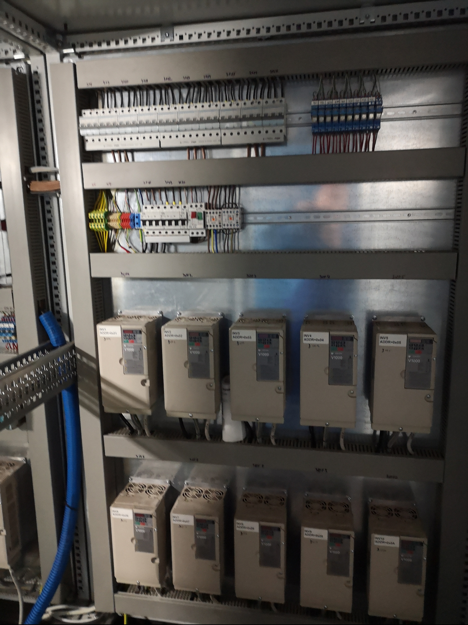 Szafa sterownicza z modułami V1000, widoczne okablowanie i listwy zaciskowe. Elementy automatyki przemysłowej w szarej obudowie, ułożone w rzędach.
