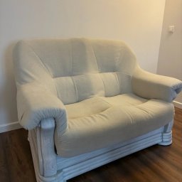 Karol Anczarski - Jasna, dwuosobowa sofa z drewnianą podstawą o żłobkowanej fakturze, ustawiona na brązowej podłodze. Widoczny napis 'po' w prawym dolnym rogu.