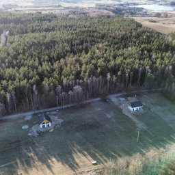 Taxi Wojciech Żuk - Szeroki kadr z drona ukazuje trzy domy jednorodzinne na skraju lasu, z polami i jeziorem w tle. Domy o prostej architekturze, z białymi ścianami i ciemnymi dachami.