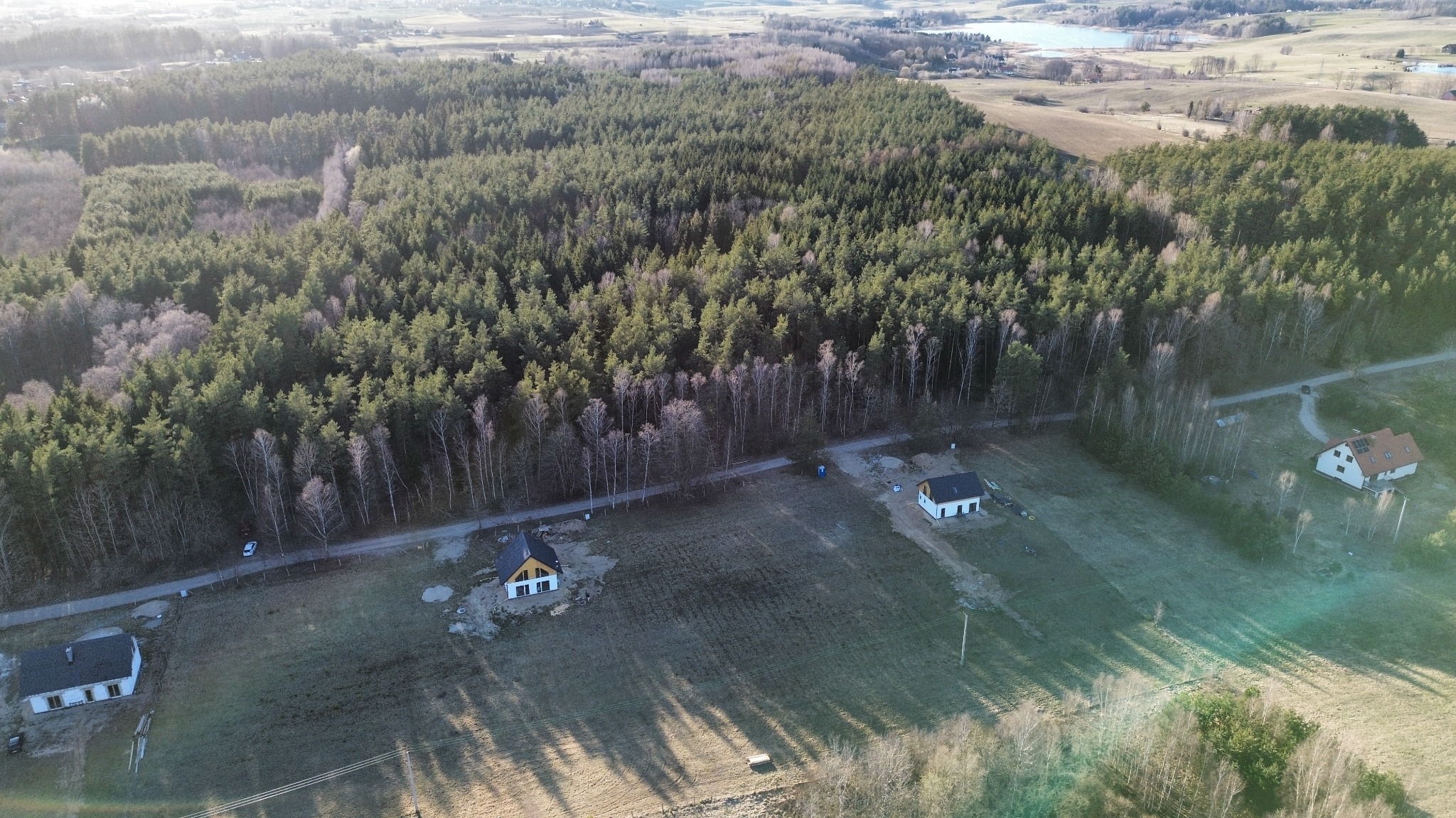 Szeroki kadr z drona ukazuje trzy domy jednorodzinne na skraju lasu, z polami i jeziorem w tle. Domy o prostej architekturze, z białymi ścianami i ciemnymi dachami.