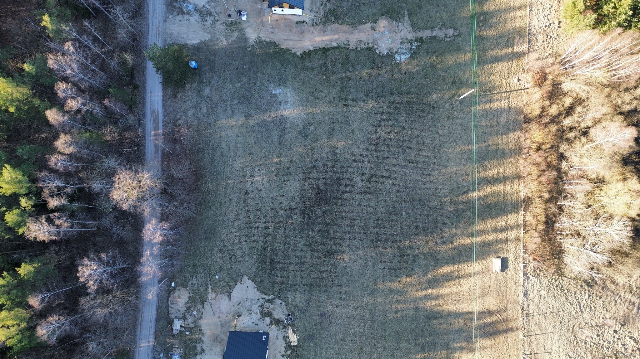 Zdjęcie z drona przedstawia rozległą działkę z widocznymi śladami po pracach ziemnych, graniczącą z lasem i drogą. W tle widoczny dom z ciemnym dachem.