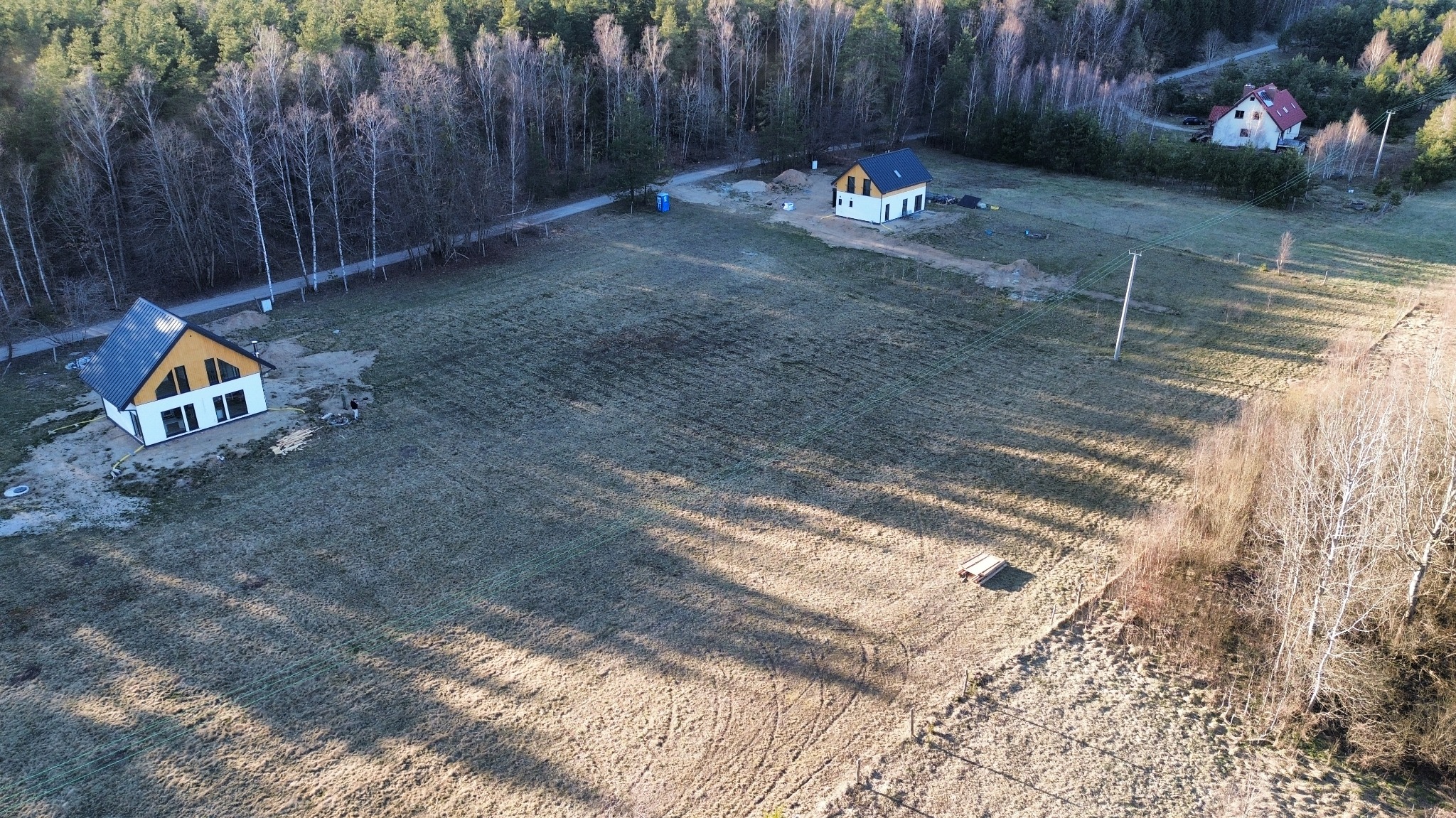 Szeroki kadr z drona ukazuje dwa domy w stanie surowym zamkniętym na rozległej działce, otoczone lasem i polami w zimowej scenerii. Widoczne ślady budowy i cień drzew.