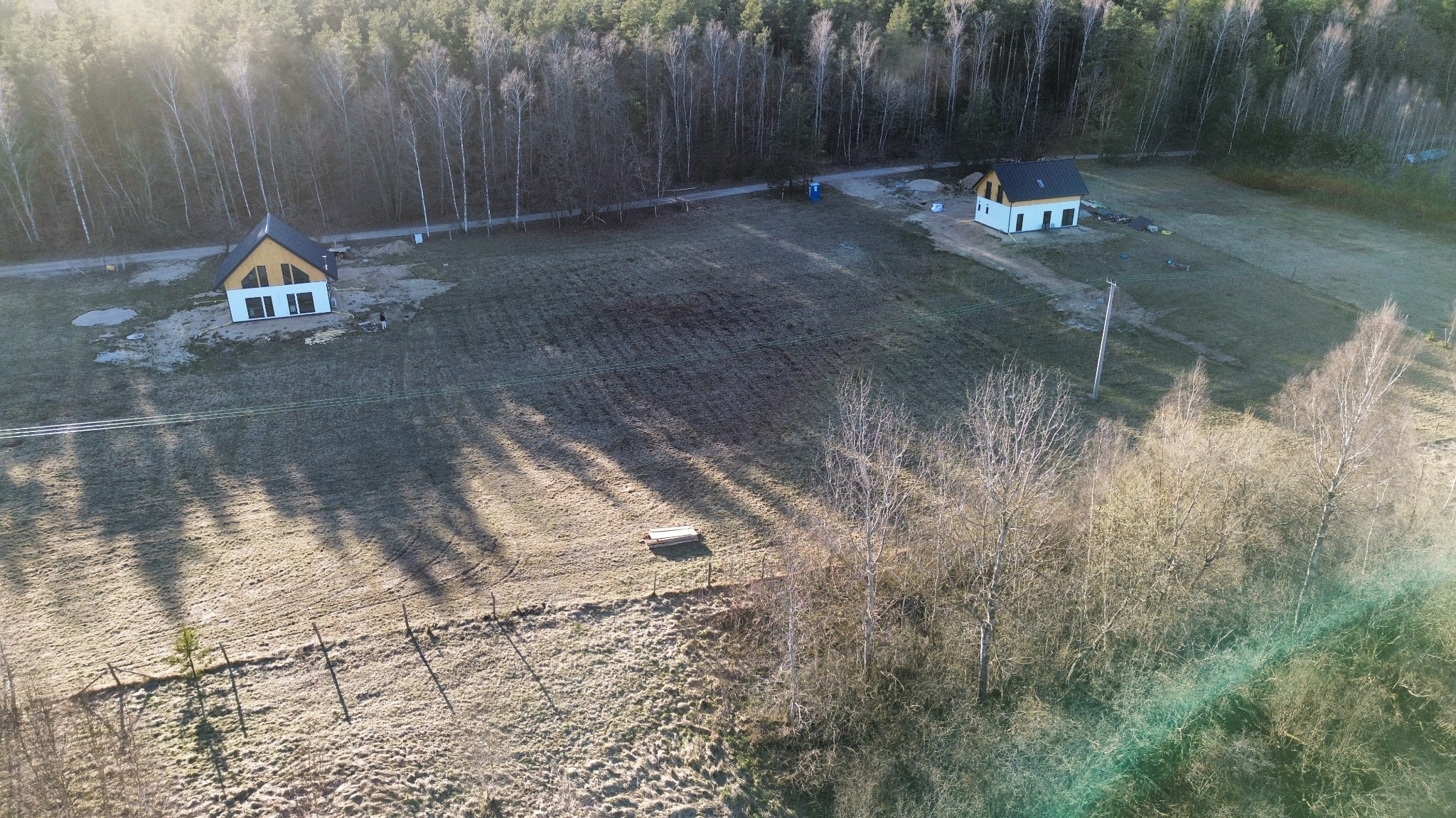 Dwa nowoczesne domy jednorodzinne z białymi elewacjami i ciemnymi dachami, widziane z lotu ptaka na tle lasu i zaoranego pola w słoneczny dzień.