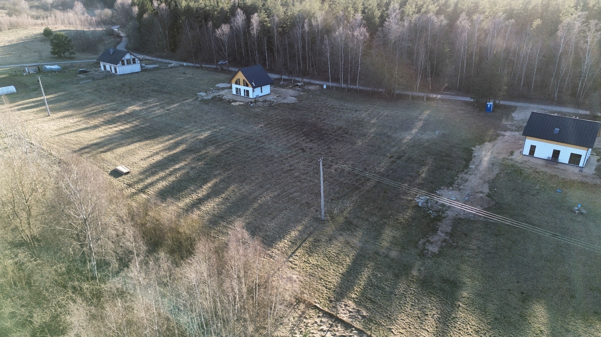 Szeroki kadr z drona ukazuje trzy domy jednorodzinne na rozległym, częściowo oświetlonym polu, z lasem w tle i słupem energetycznym na pierwszym planie.