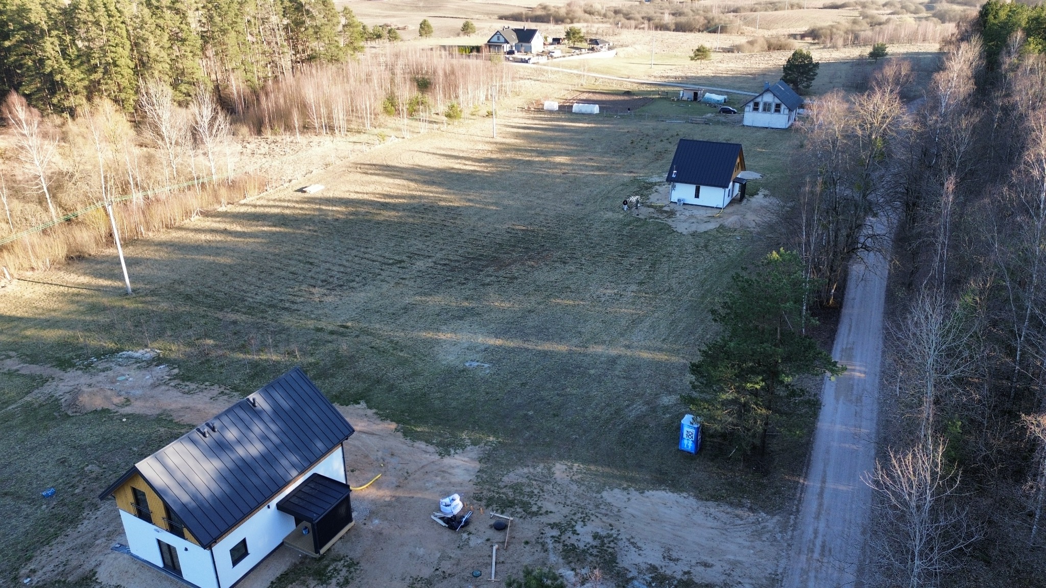 Zdjęcie z drona przedstawia kilka domów w różnej fazie budowy na rozległym, trawiastym terenie z drzewami. Jeden z domów jest prawie ukończony, z czarnym dachem i białymi ścianami.