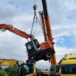 Nokop Andrzej Filipiak - Pomarańczowa koparka Hitachi ZA 16 podnoszona przez dźwig na tle pochmurnego nieba, z widocznymi innymi maszynami budowlanymi w tle.