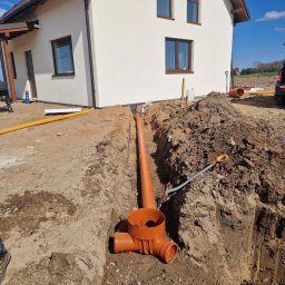 Nokop Andrzej Filipiak - Pomarańczowe rury kanalizacyjne ułożone w wykopie ziemnym obok jasnego domu z brązowymi oknami, widoczne narzędzia i żółta rura drenażowa. Słoneczny dzień.