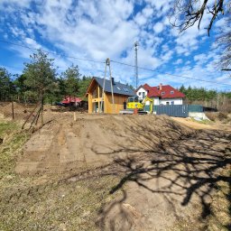 Nokop Andrzej Filipiak - Żółta koparka na wzgórzu z przygotowanym terenem pod budowę domu. W tle domy, drzewa i błękitne niebo z chmurami. Cień drzewa w dolnej części kadru.